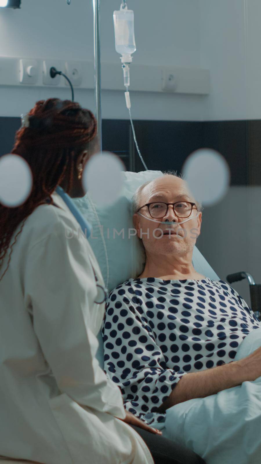 Nurse and african american doctor tending to ill patient in hospital ward bed at medical facility. Sick old man with nasal oxygen tube and IV drip bag recovering from injury and disease