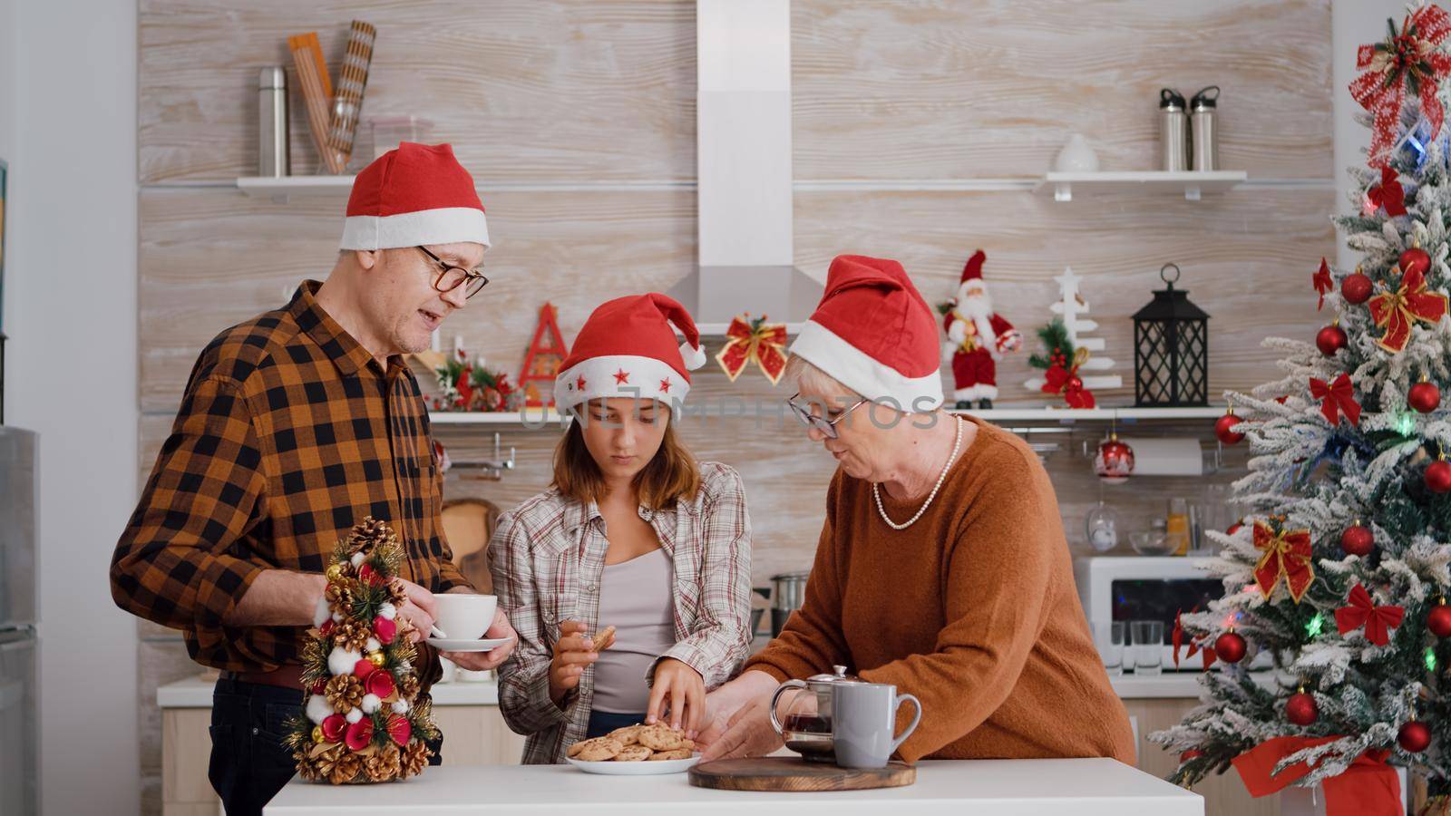 Family with santa hat speding christmas holiday together enjoying christmastime smiling in decorated kitchen. Grandparents drinking coffee while grandchild eating delicious chocolate baked cookies