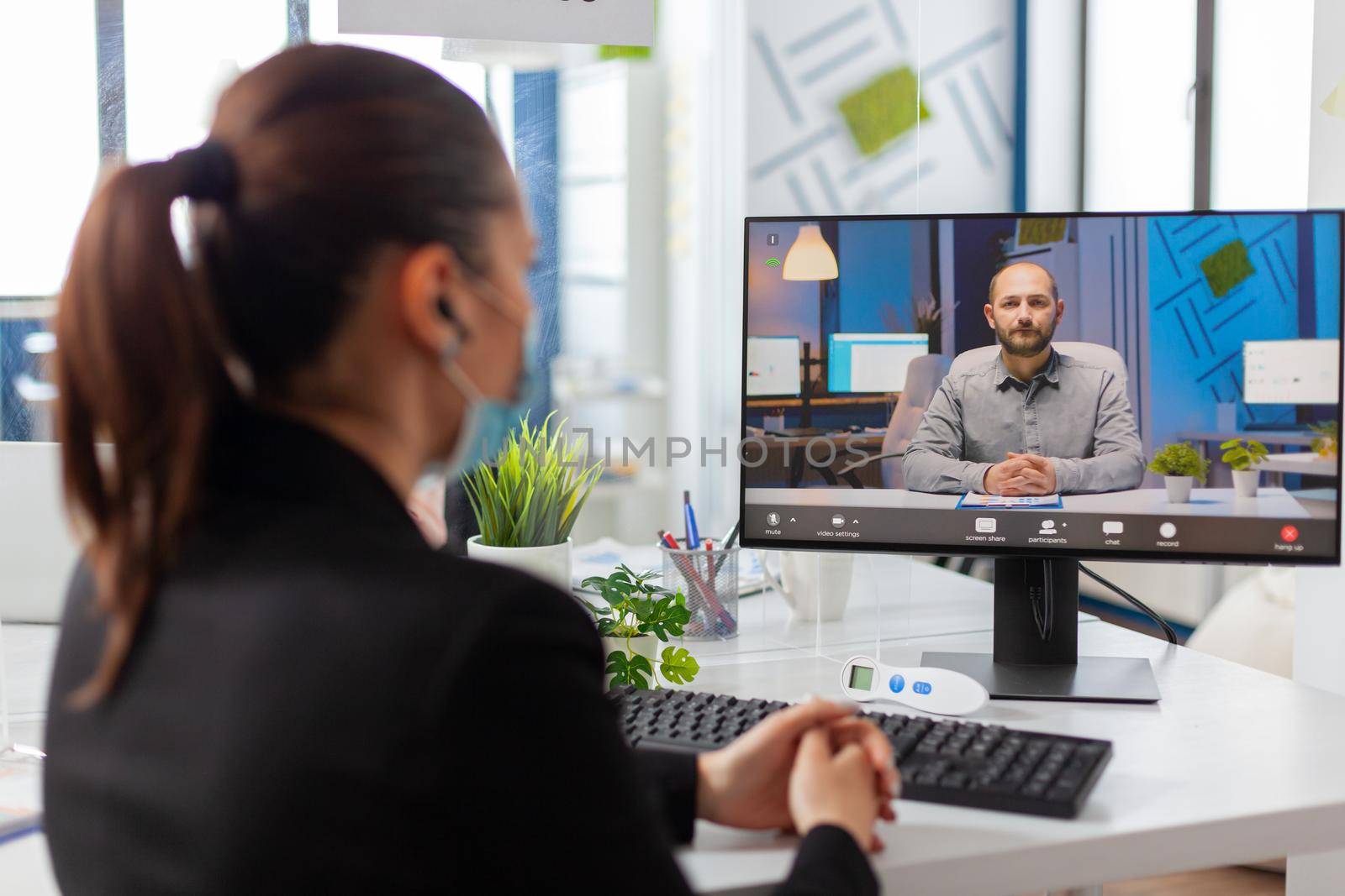 Woman entrepreneur talking with partner on remote call in workplace during global pandemic with coronavirus, wearing face mask as safety prevention in workplace.