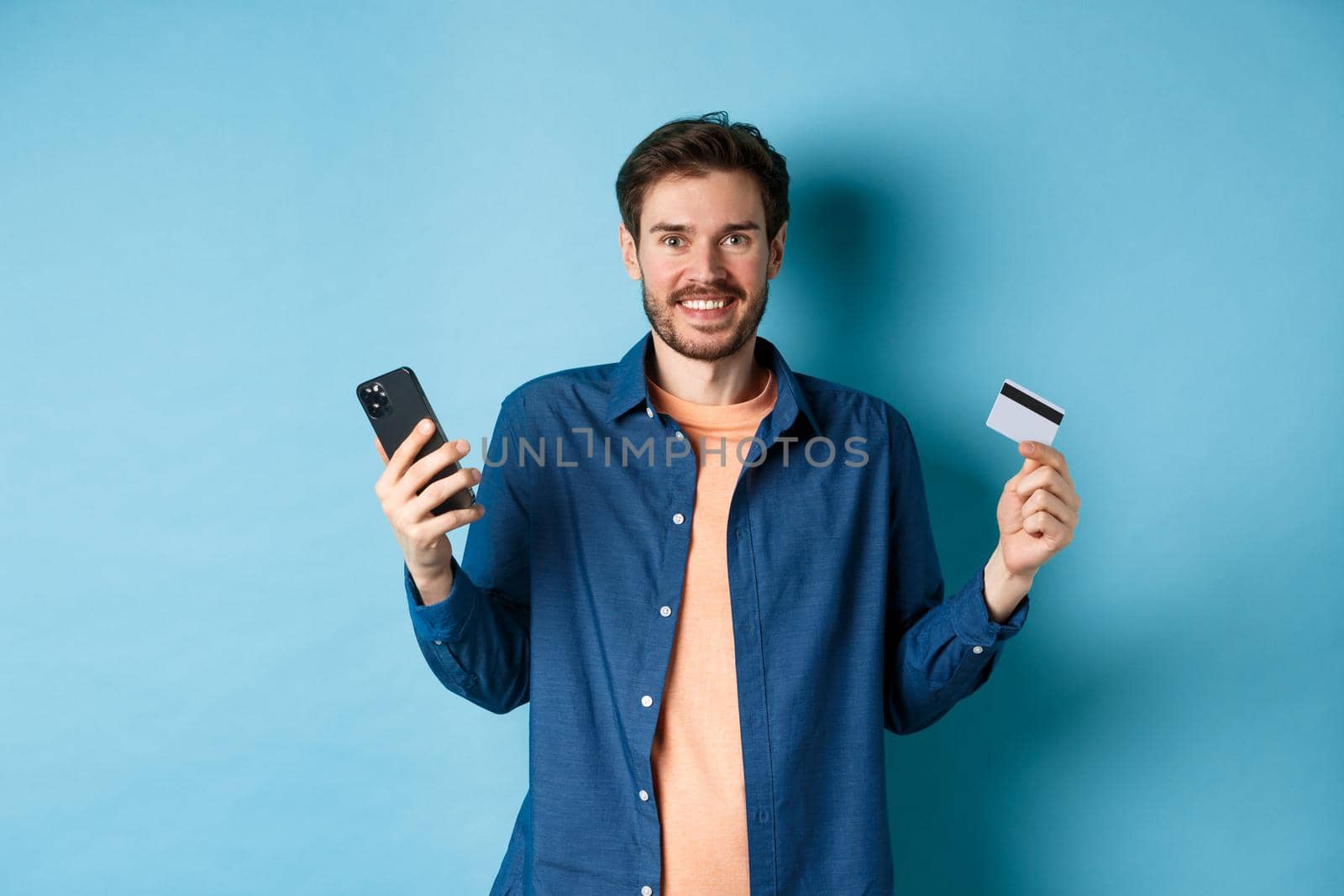 E-commerce concept. Excited young man holding smartphone and plastic credit card, shopping online, standing on blue background by Benzoix