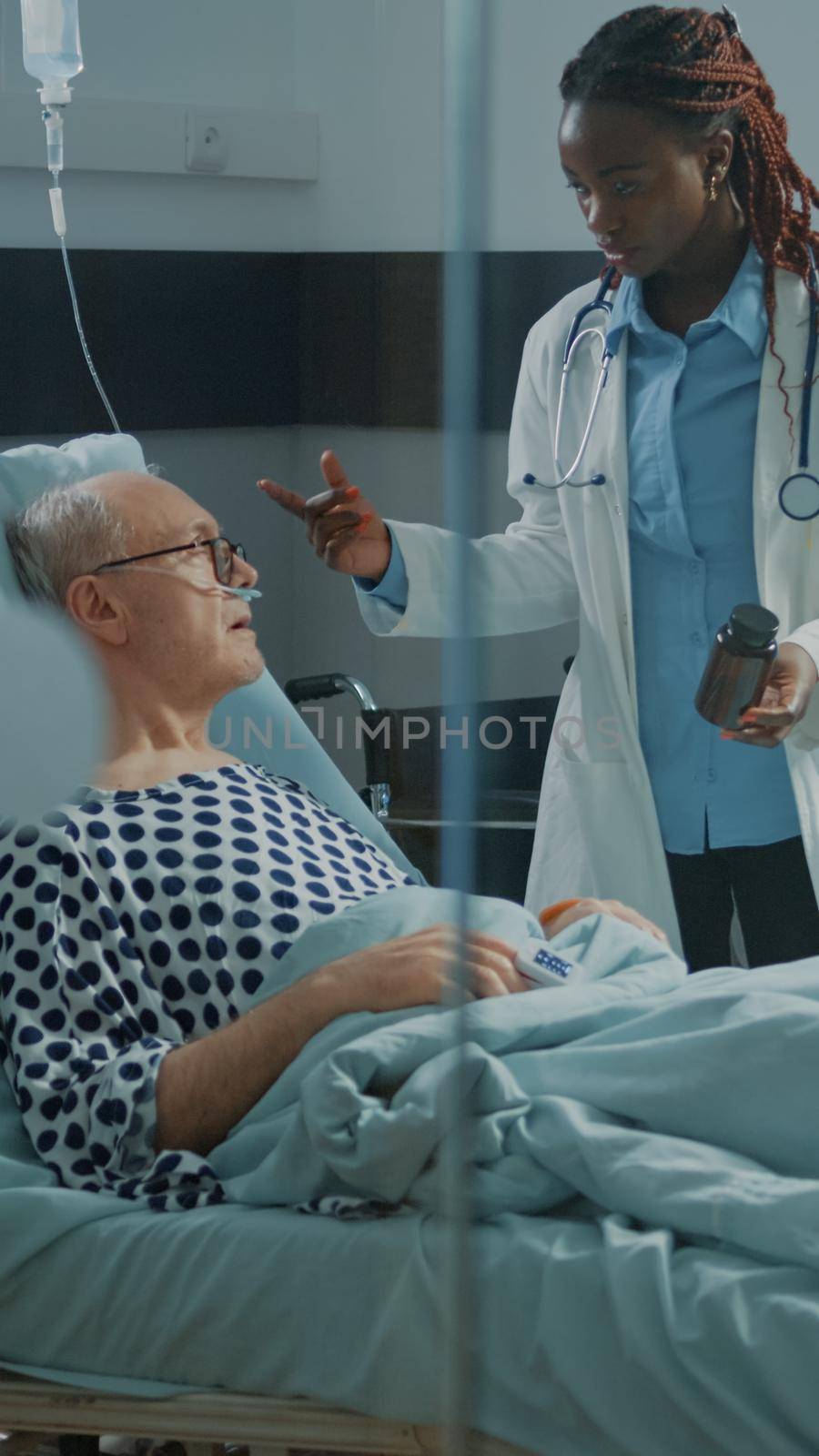 Sick patient laying in hospital ward bed at facility waiting for african american doctor to give bottle of pills as medical treatment. Medic curing old man with health problems at clinic