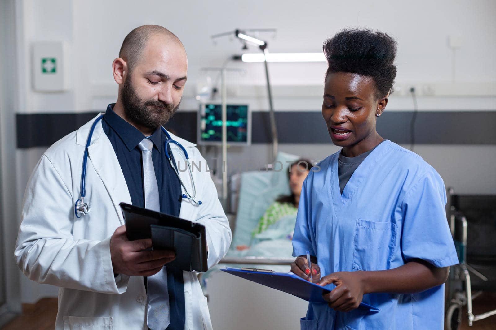 Man practitioner discussing with black assistant in hospital ward during medical consultation. Sick woman patient resting in bed while physician doctor monitoring illness disease