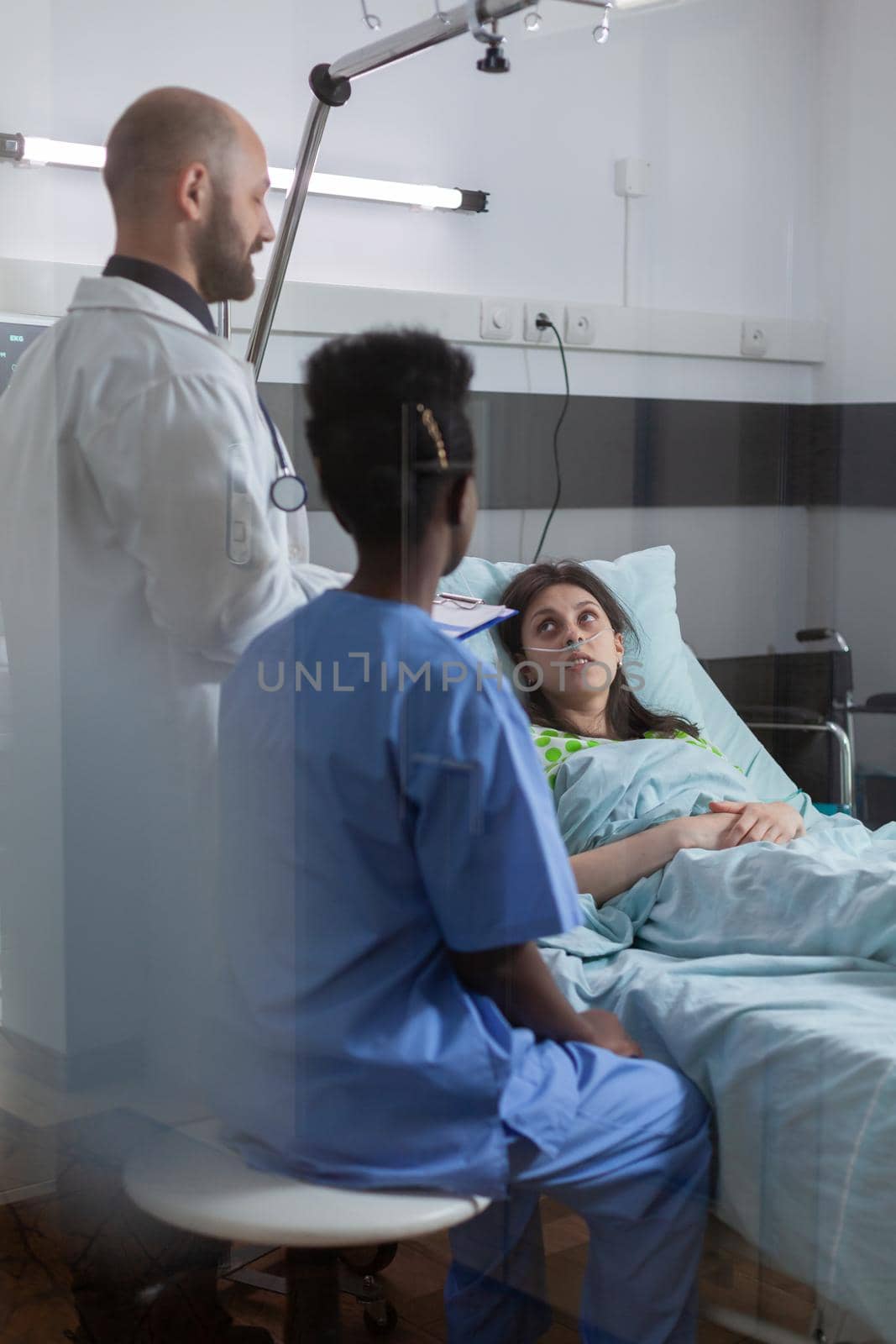 Medical team monitoring sick woman explaining sickness symptom examining medical recovery in hospital ward. Patient resting in bed while doctor writing treatment during rehabilitation appointment
