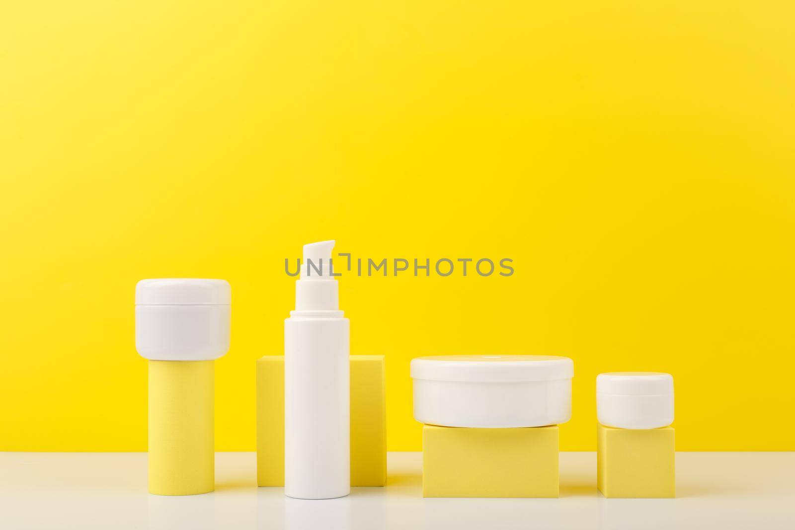 Trendy colorful composition with set of cosmetic bottles with beauty products on geometric props against yellow background with copy space. Concept of creams with citrus extracts or skin products with sunblock