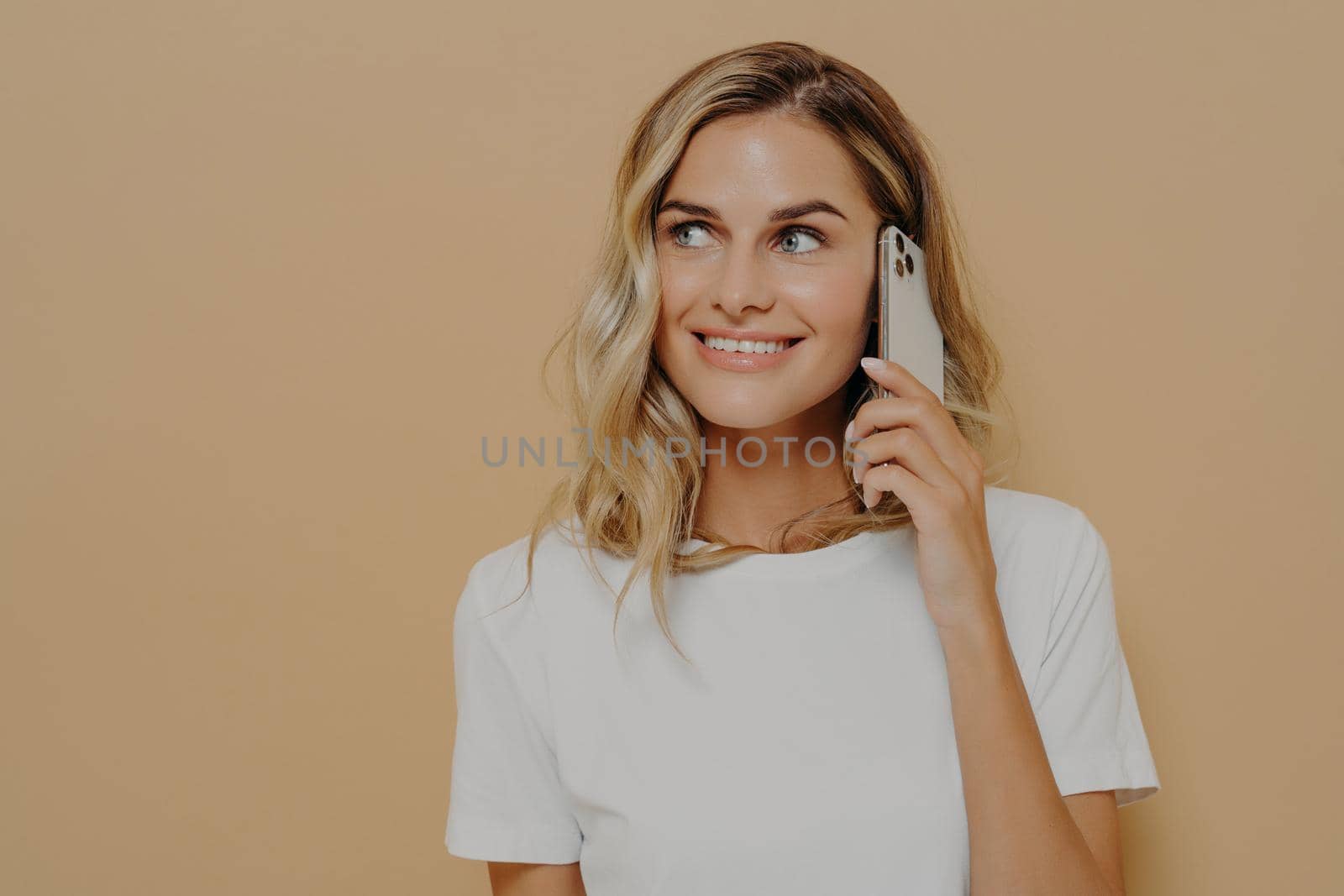 Young blonde smiling woman using cell phone and looking aside, enjoying pleasant conversation with her boyfriend, dressed in white t shirt. Modern technologies and communication concept