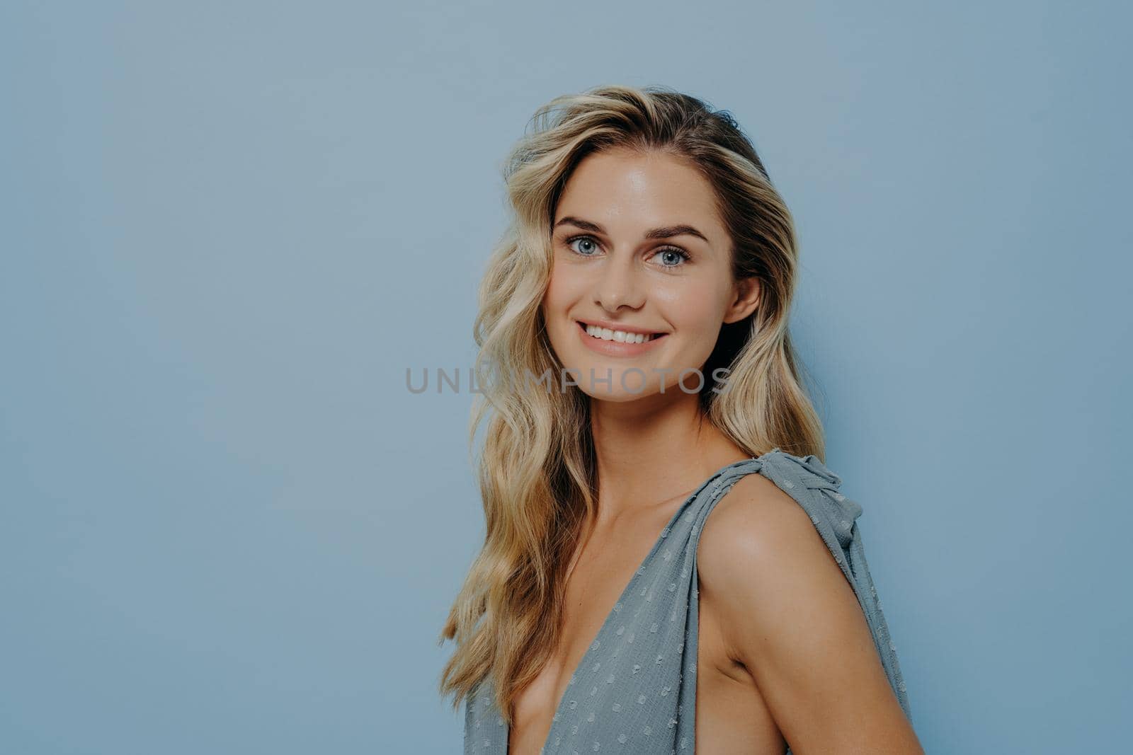 Portrait of cheerful smiling blonde girl dressed in blue dress, standing sideways with toothy smile while looking at camera, calm and relaxed at same time, posing in front of blue background