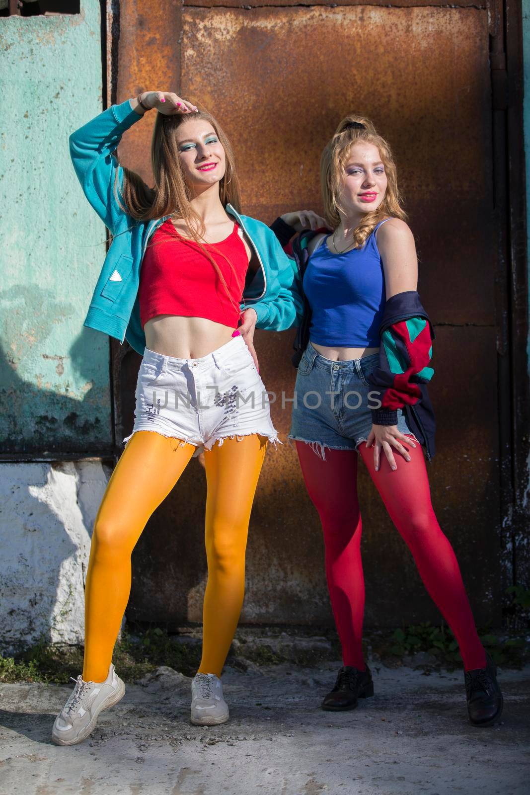 Two funny girls in bright clothes in the style of the nineties are standing against the background of a rusty door. Russian village girls.