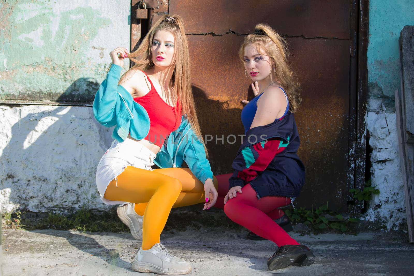 Two funny girls in bright clothes in the style of the nineties are sitting against the background of a rusty door. Russian village girls.