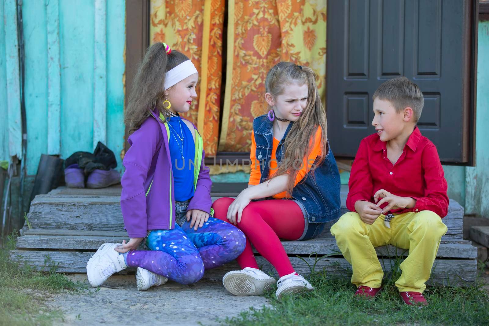 Funny little children: girls with bright makeup dressed in the style of the nineties and a boy in a red shirt are sitting on the village porch of the house. Russian village children.