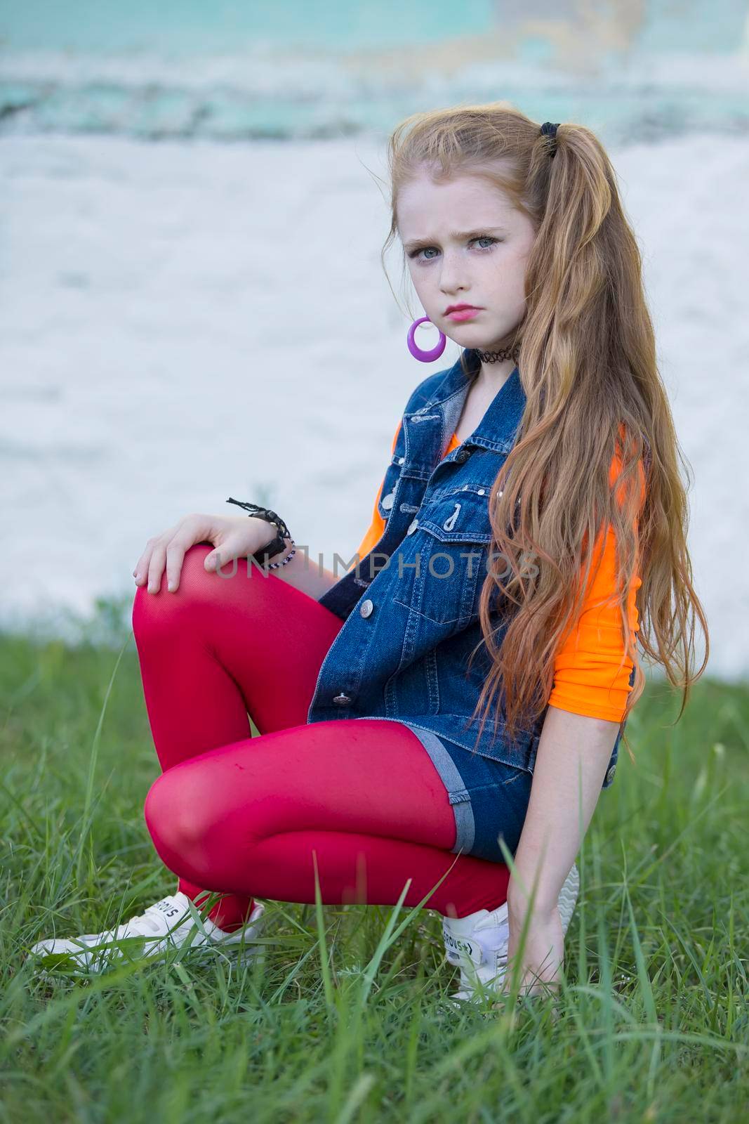 Portrait of a little red-haired girl with bright makeup in a denim vest and red leggings. by Sviatlana