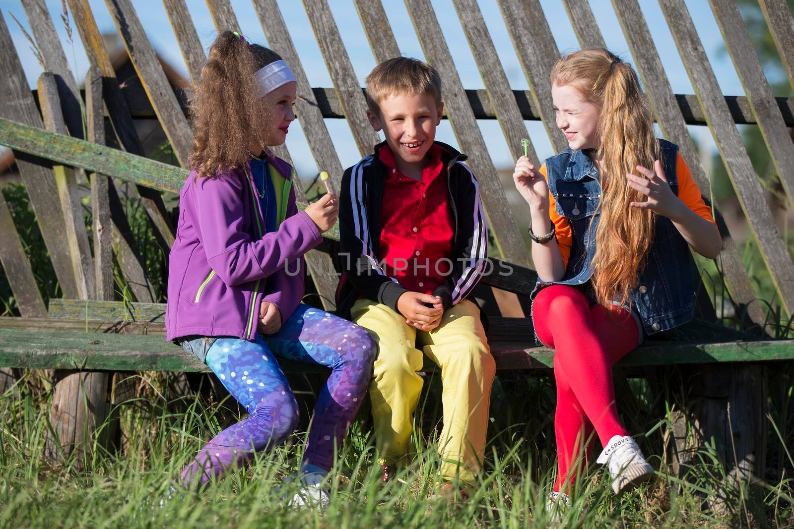 Funny little children: girls with bright makeup dressed in the style of the nineties and a boy in a red shirt are sitting on a bench by a skewed fence. Russian village children. by Sviatlana