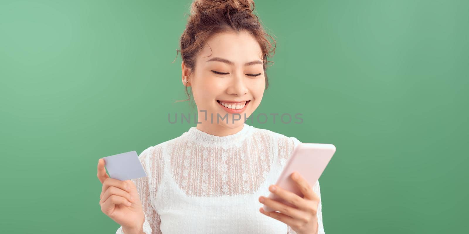 A beautiful portrait of a happy young girl showing plastic credit card while holding mobile phone isolated over green background