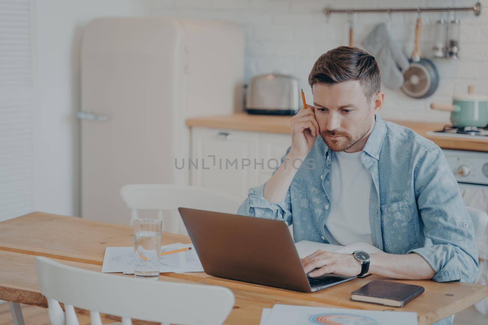 Serious focused determined young bearded businessman or freelancer in casual clothes sitting at kitchen table and typing on laptop, working on project online, researching or writing email to client