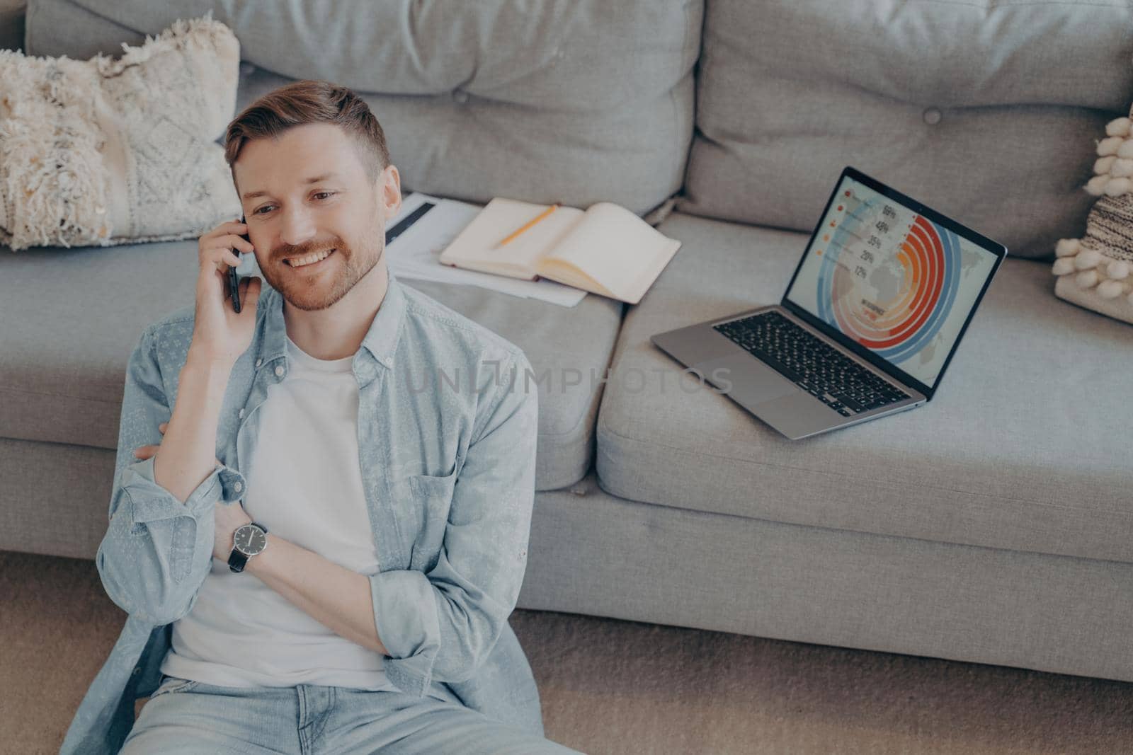 Happy young man calling family to inform about receiving promotion at his company after submitting successful project idea, sitting on floor while resting against couch with open laptop showing graphs