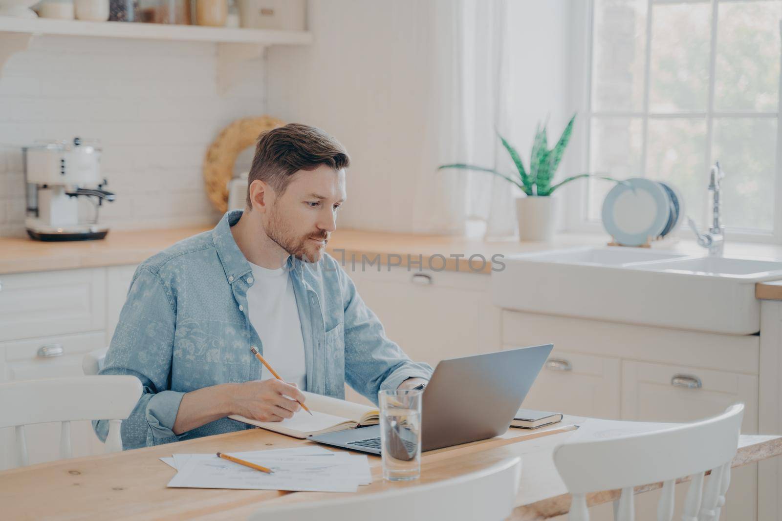 Portrait of concentrated male freelancer working on project remotely from home while sitting at kitchen table with laptop, wears casual clothes. Business online and freelance concept