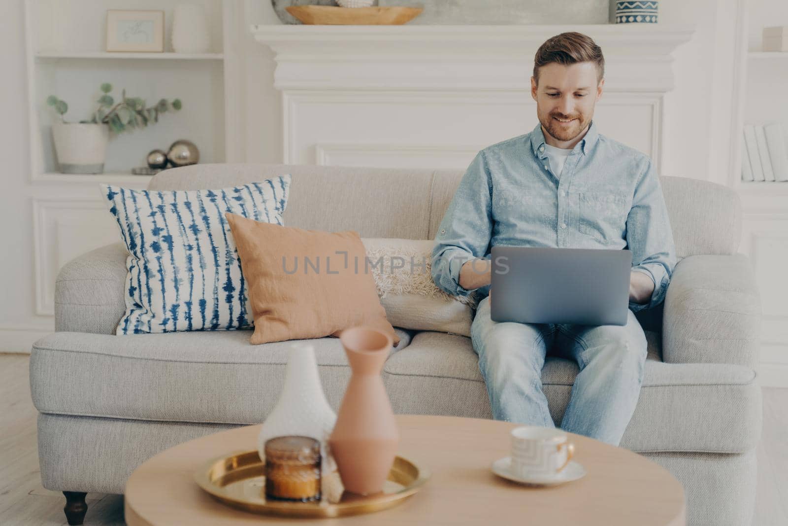 Young positive male freelancer working on his commissioned project using notebook, sitting alone on couch with lots of different pillows in living room, table with cup of coffee next to him