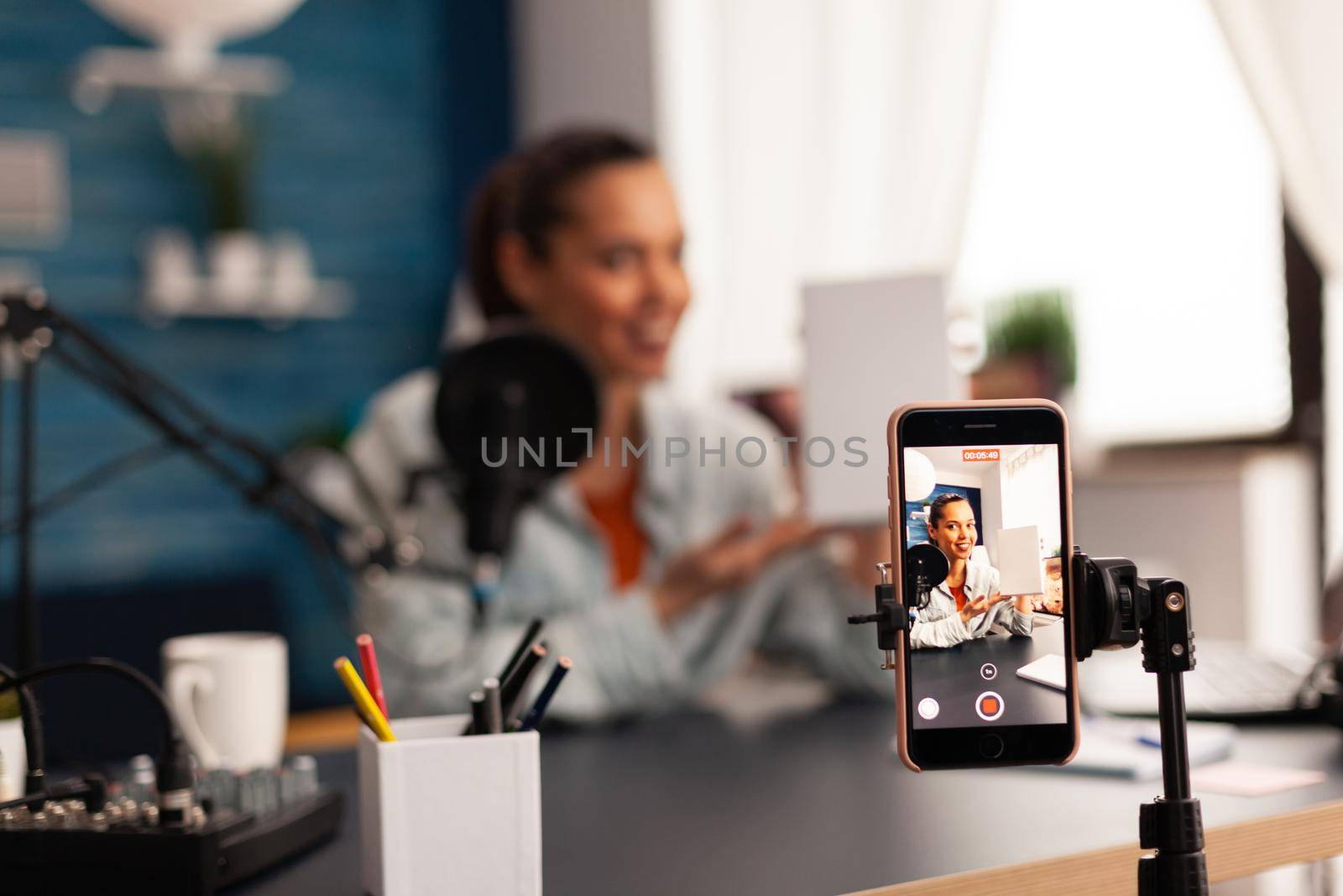 Influencer holding white gift box while recording videoblog for social media. Creative content creator making broadcast concept speaking and looking at smartphone on tripod at home studio podcast