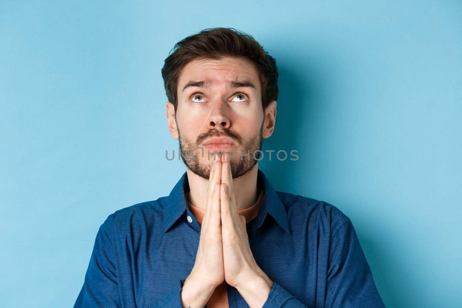 Close up shot of hopeful guy pleading God, holding hands in begging gesture and looking up in sky, standing on blue background supplicating.