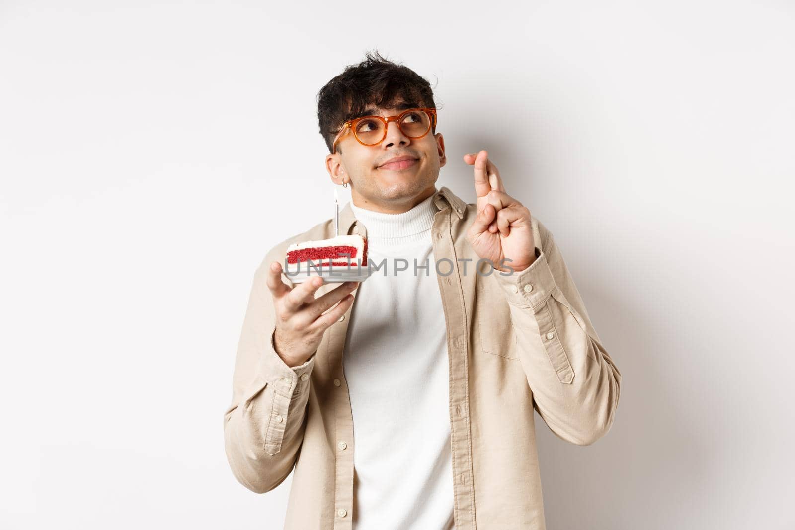 Cute hopeful guy in glasses making birthday wish, holding cake and fingers crossed, looking up and praying, hope dream come true, standing on white background.