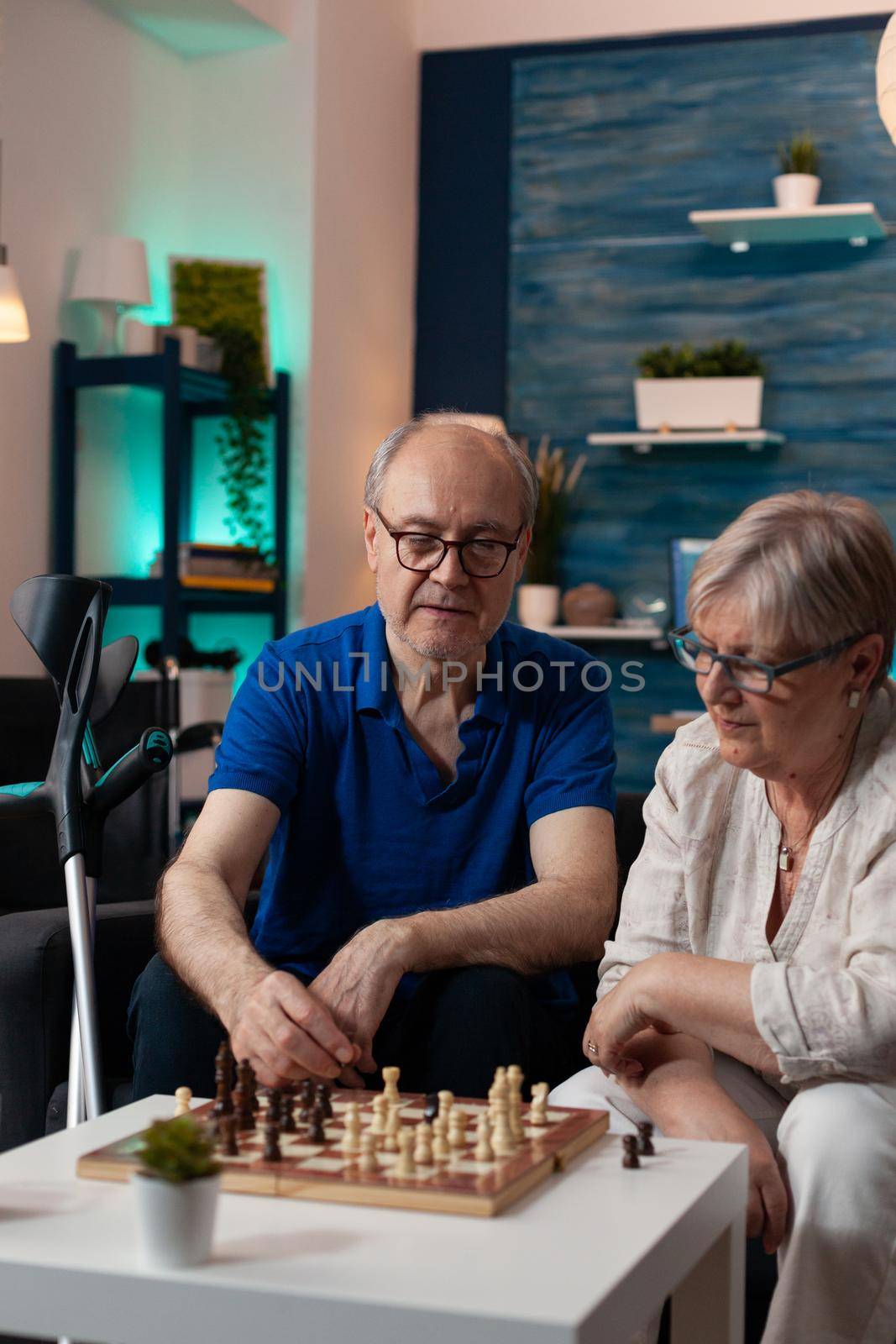 Retirement aged people sitting on living room sofa at home while playing chess game on board to relax. Caucasian old couple enjoying indoor fun activity resting on couch with crutches