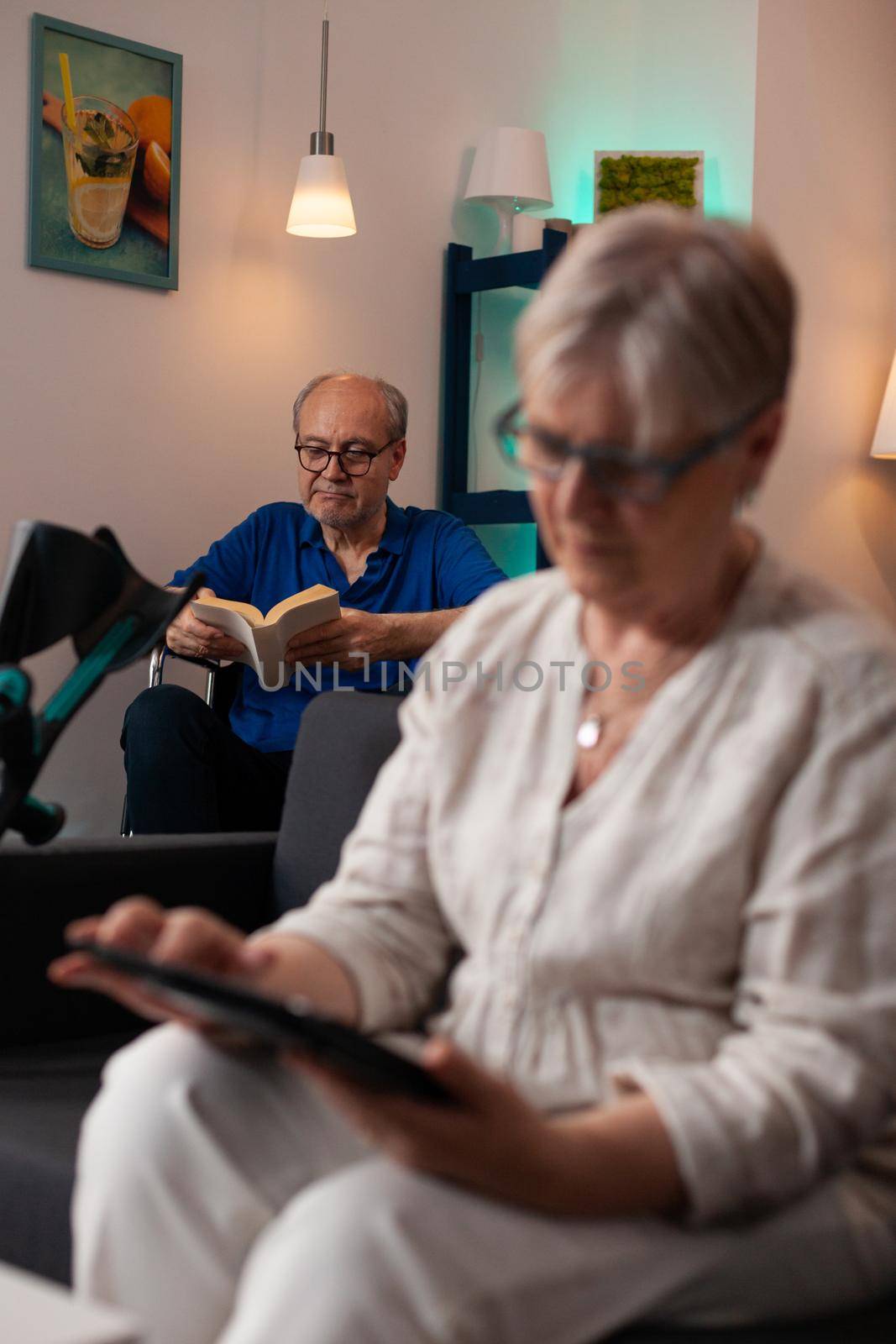 Senior retired woman looking at digital tablet gadget sitting on couch in living room while old man in background reading book. Elder couple with disability problems relaxing at home