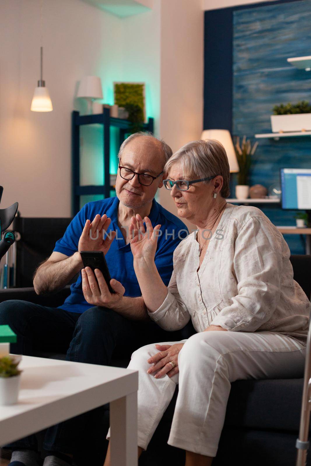 Elderly couple waving at video call webcam using smartphone technology with modern online internet connection sitting on couch. Caucasian people meeting on virtual conference with family