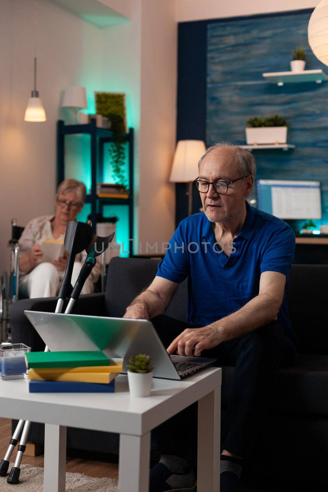 Handicap retired people relaxing at modern home with technology and medical equipment. Old man using digital laptop computer device sitting on sofa with crutches and woman in wheelchair