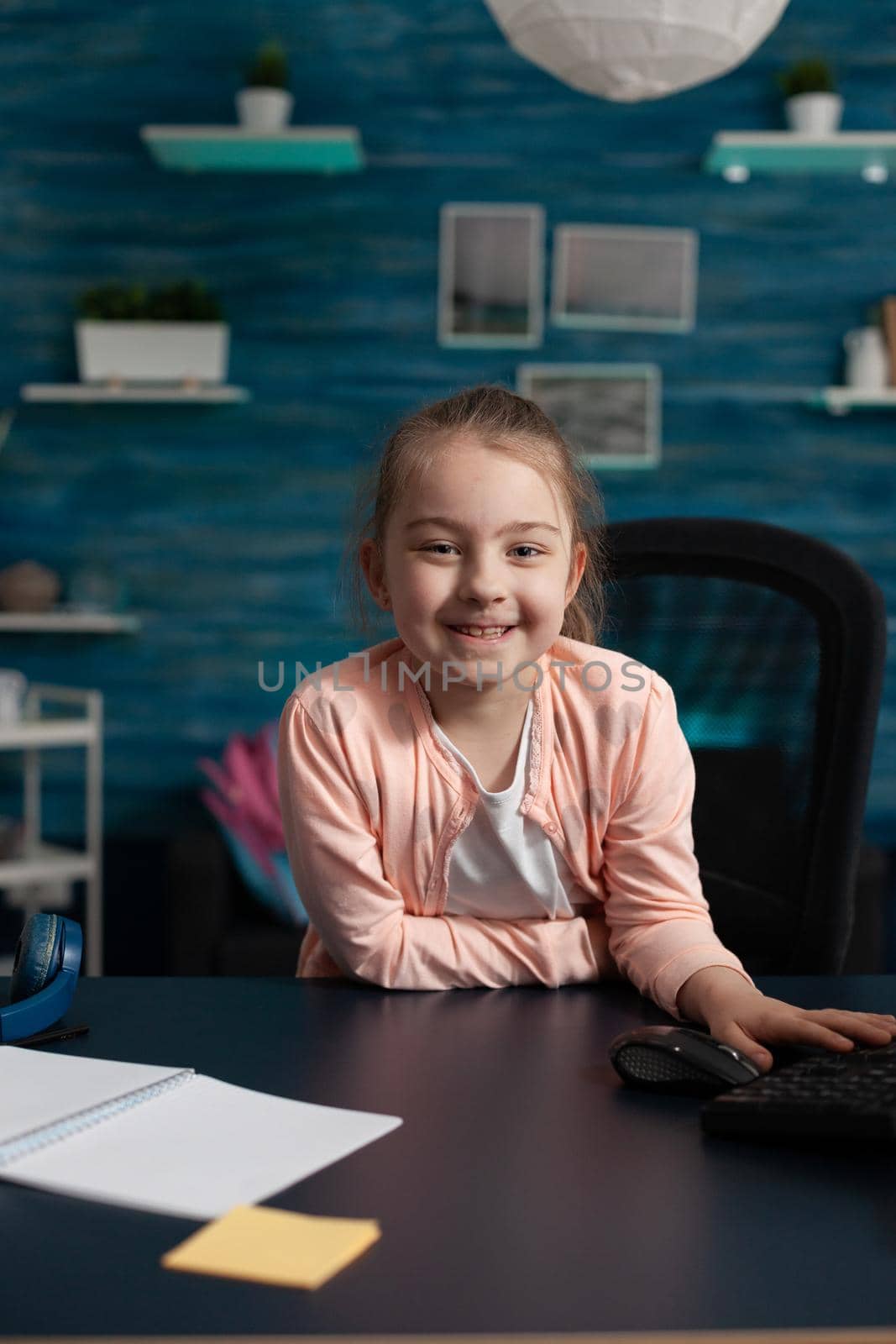 Joyful little girl smiling sitting at desk for online school work on internet classroom lecture. Clever child with educational supplies notebook pen preparing for communication meeting
