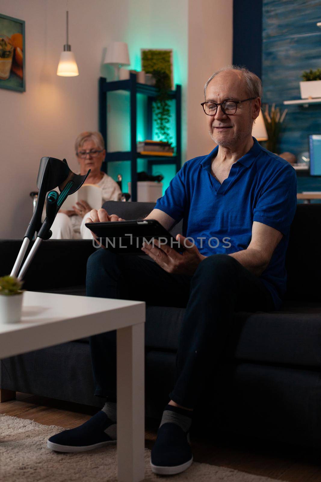 Couple of pensioners sitting at home with modern technology and healthcare support equipment. Caucasian man using digital tablet gadget on couch with crutches and woman in wheelchair