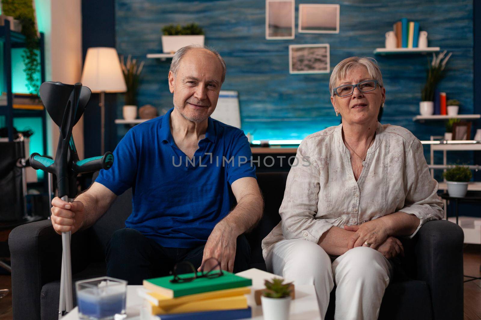 Portrait of elderly couple sitting together at home in decorated living room. Caucasian old husband with transportation disability crutch and senior wife smiling looking at camera