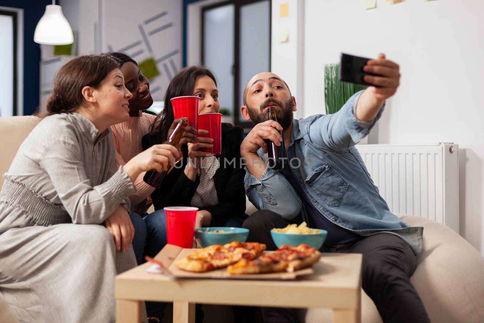 Diverrse group of coworkers taking selfie on smartphone after work at office party. Cheerful colleagues having fun at celebration meting with pizza chips bottles and cups of beer alcohol