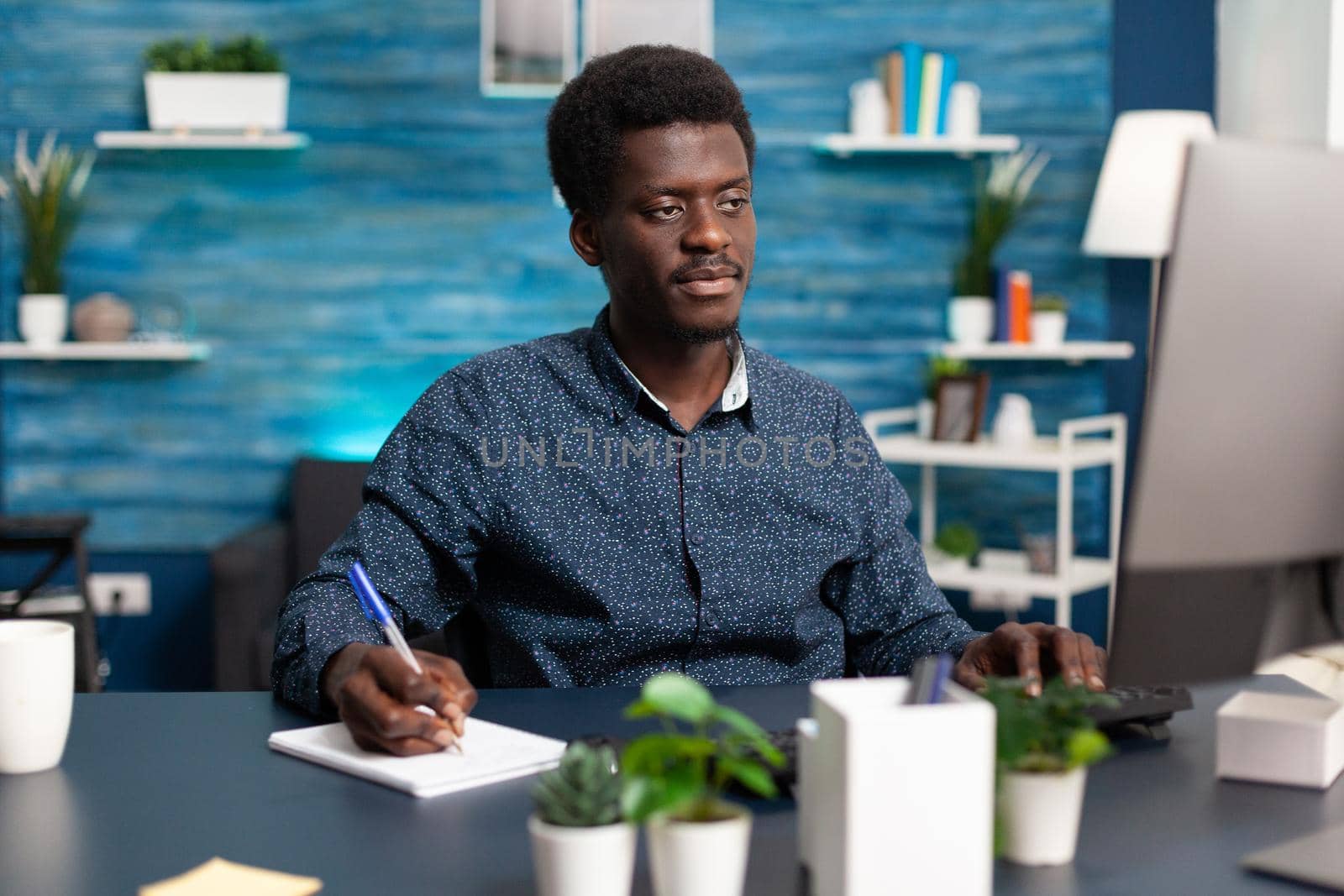 Freelancer taking notes on notebook looking at computer on home office desk. African american man remote working from home and doing online college courses for social media business