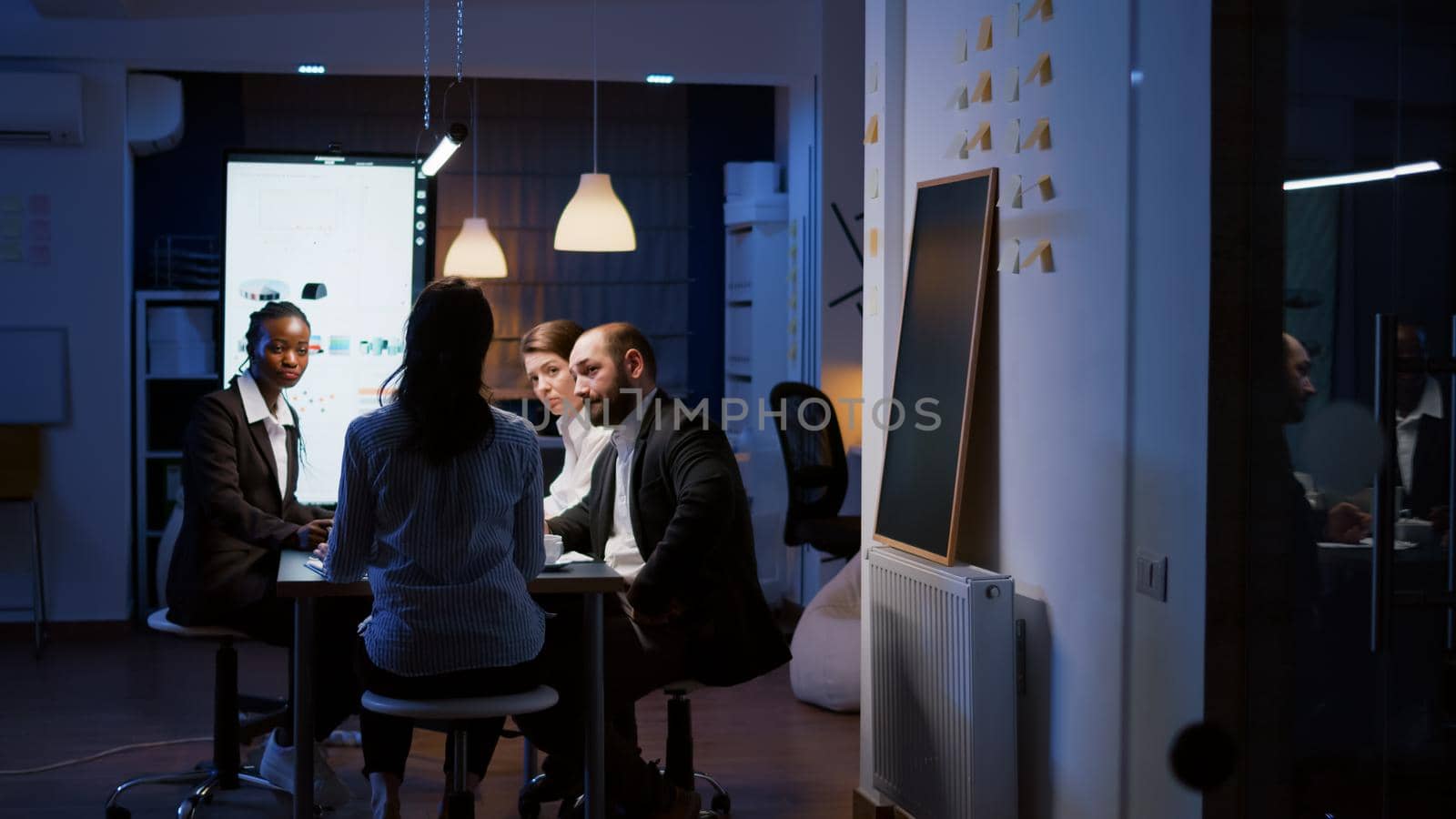 Selective focus of workaholic businesswoman back discussing financial graphs analyzing company paperwork overworking in office meeting room late at night. Multi-ethnic coworkes brainstorming ideas