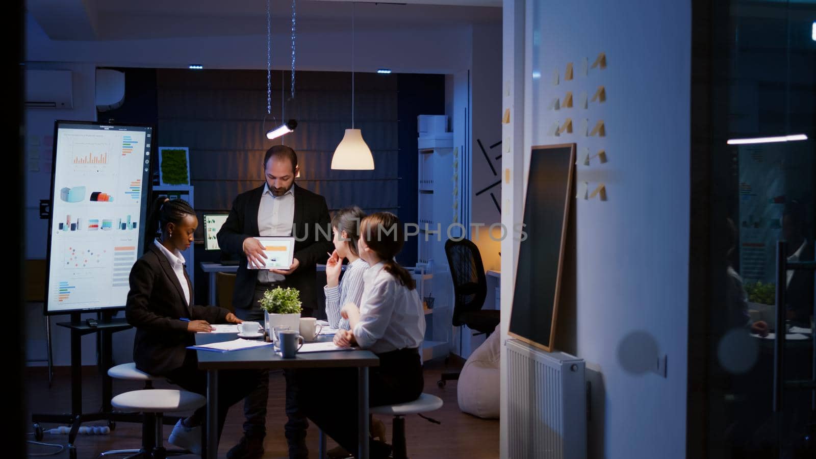 Workaholic overworked businessman explaining company strategy using tablet for presentation working overtime in meeting office room late at night. Diverse multi-ethnic teamwork brainstorming ideas