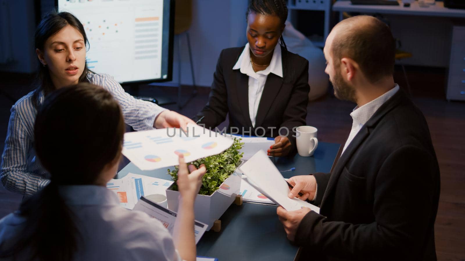 Diverse multi ethnic businesspeople sharing management presentation on paperwork discussing company strategy. Teamwork brainstorming ideas while working in meeting room late at night