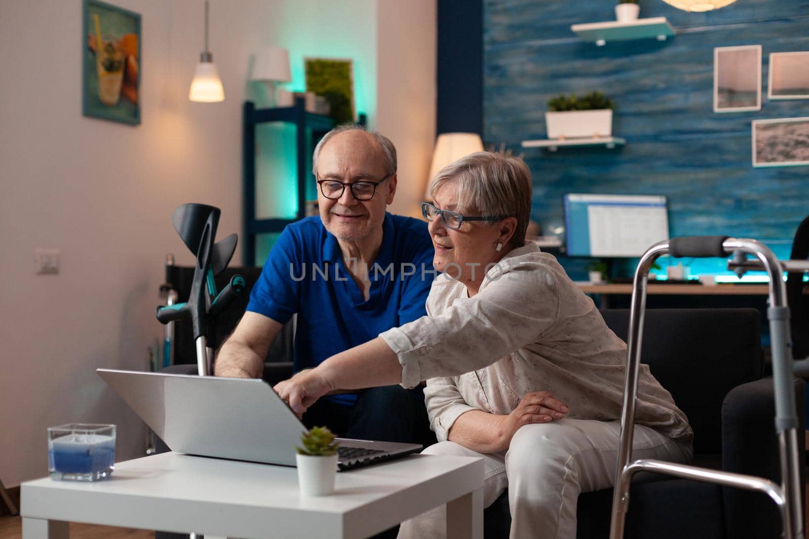 Gray haired woman old man analyzing laptop screen computer monitor at home on sofa. Retired caucasian couple using modern technology device having walk frame crutches and wheelchair