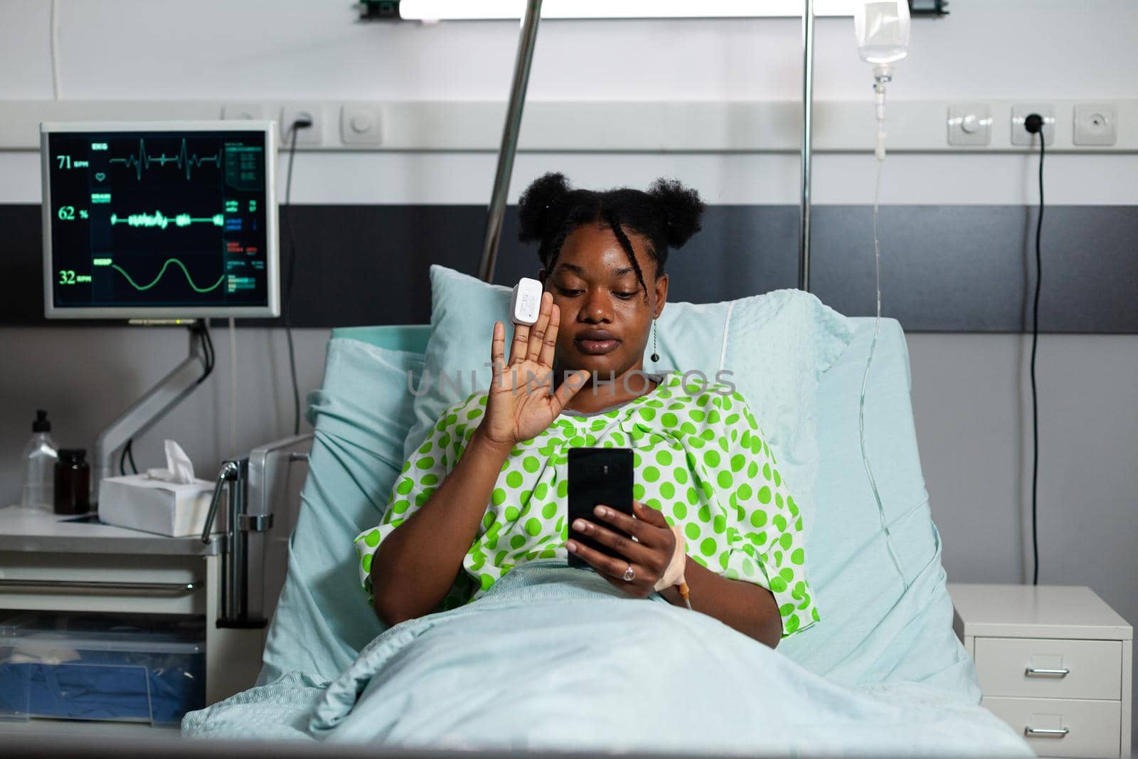 Sick african american patient using video call technology on smartphone recovering in hospital ward bed. Young person with disease waving while being on online internet conference with family