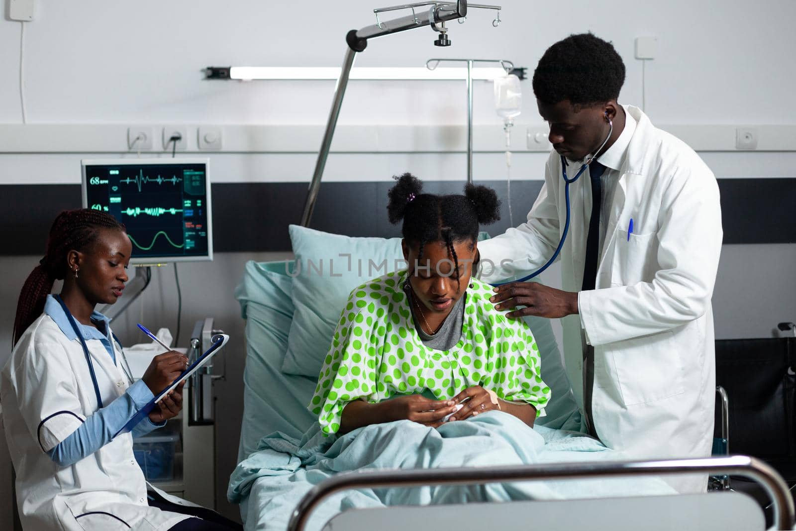 African american doctor consulting young patient in hospital ward bed using stethoscope for diagnosis. Woman writing results, medical team working on treatment, medicine for girl
