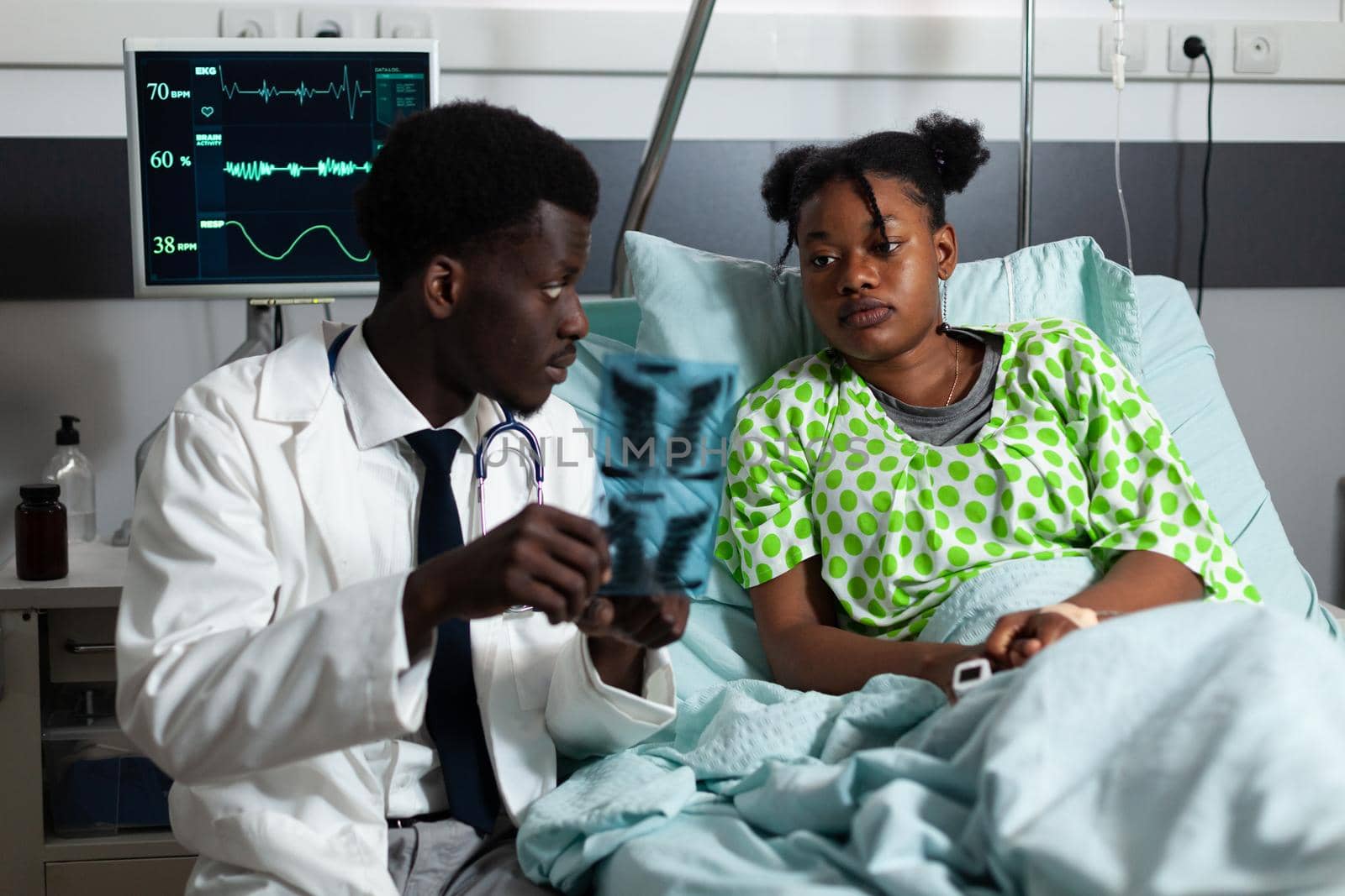 African american doctor and patient looking at x ray in hospital ward at healthcare clinic. Afro man holding scan for treatment development and medicine. Young adult with disease sitting in bed