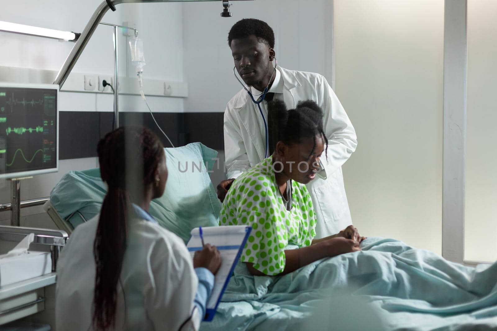 Team of african ethnicity doctors examining patient in hospital ward sitting on bed. Man using stethoscope for consultation while woman assisting girl with disease, sickness, illness