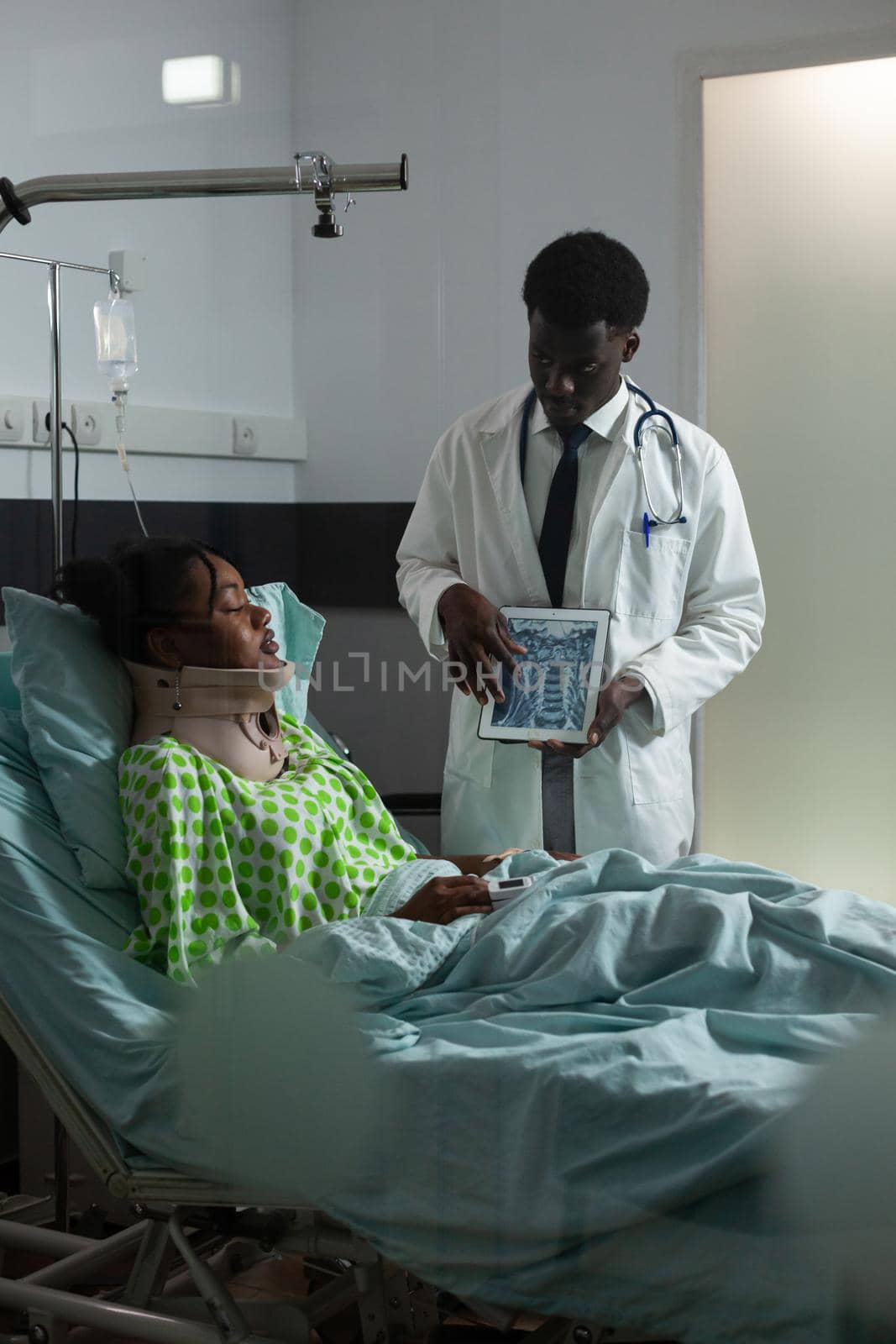 Doctor of african ethnicity showing x ray results to young patient laying in hospital ward bed with cervical neck collar for support and healthcare. Man and sick girl looking at tablet