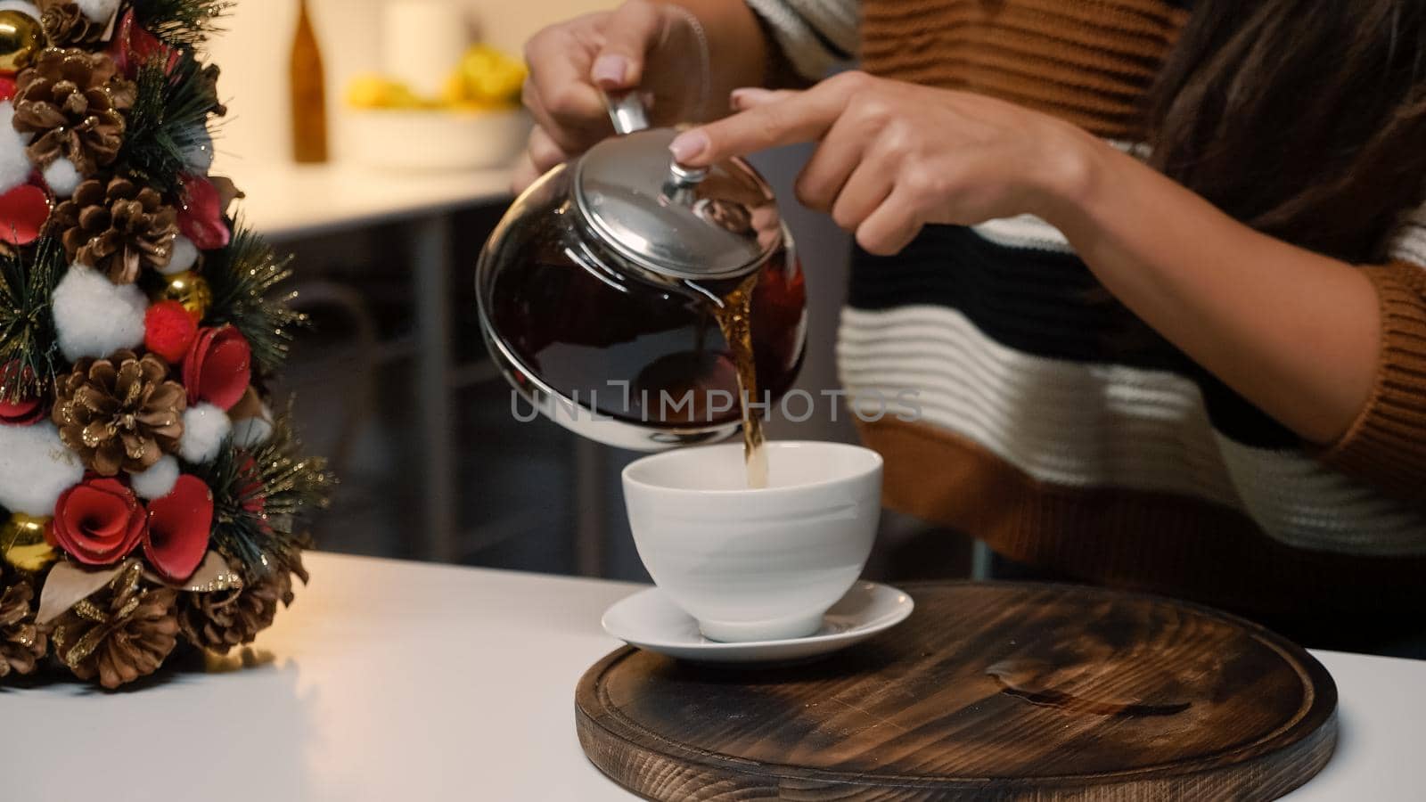 Festive young woman pouring tea from kettle in cup at home with christmas decorations and ornaments. Close up of kitchen utensils on counter while preparing for holiday festivity