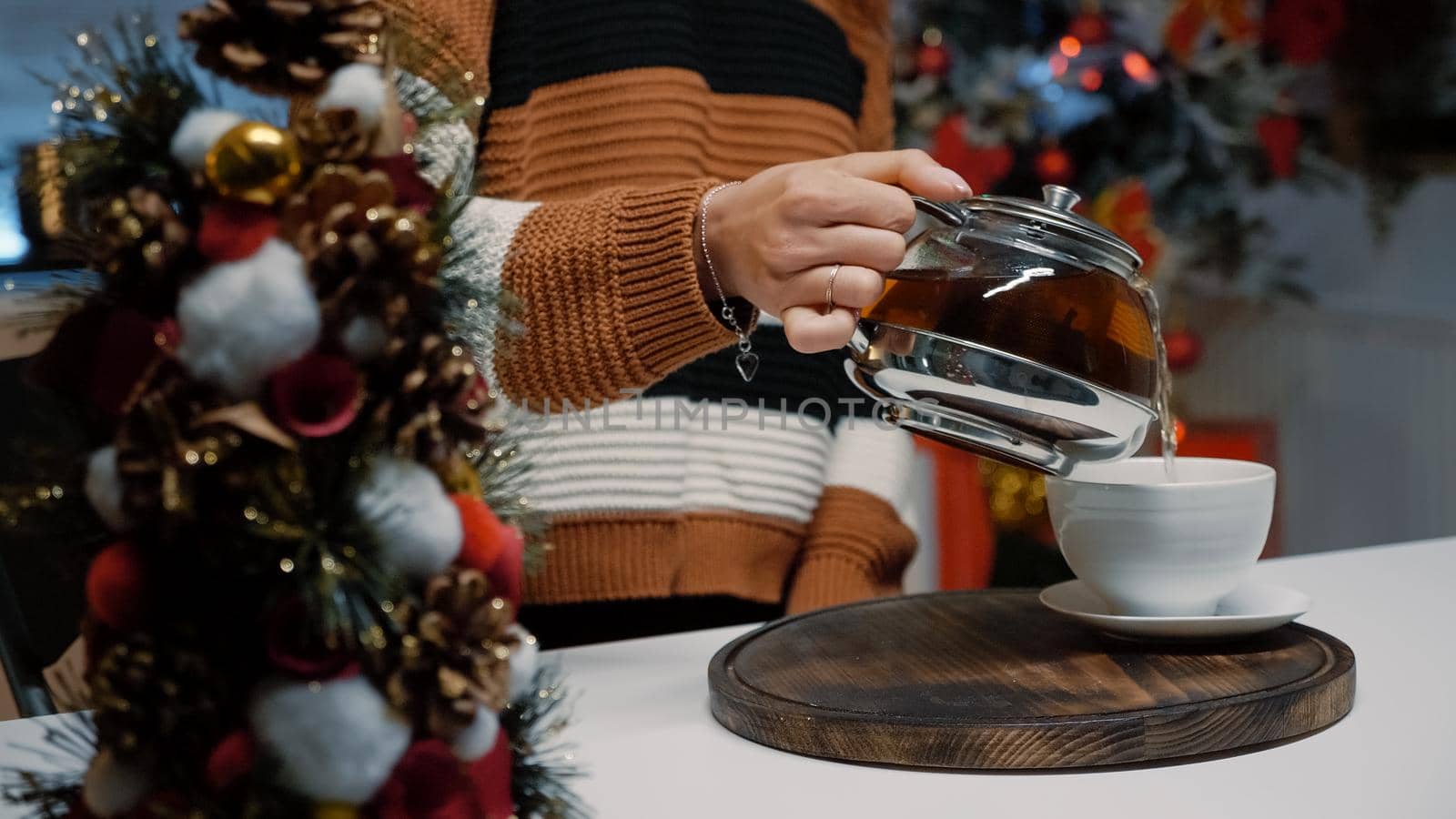 Close up of woman pouring cup of tea from kettle at home in kitchen decorated for christmas season. Counter with winter decorations and mug. Young person preparing hot drink for warmth