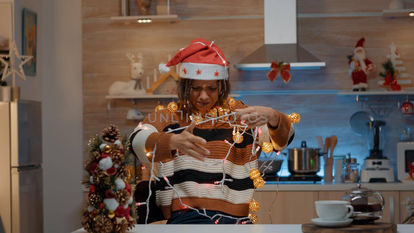 Woman decorating kitchen with festive ornaments while getting tangled in christmas string lights for tree. Young person holding illuminated garland for holiday celebration preparations