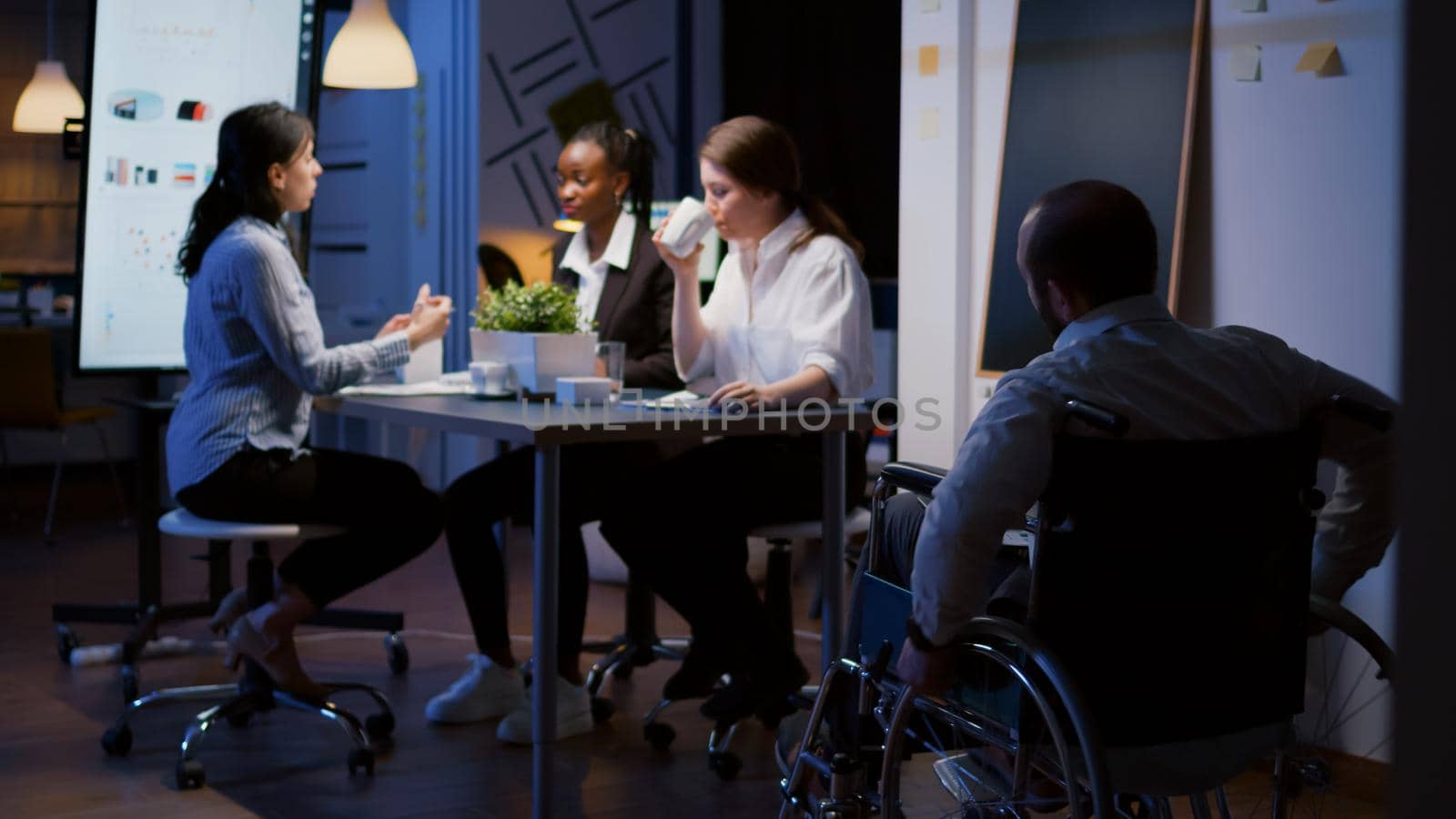Focused overworked handicapped businessman in wheelchair sharing financial paperwork statistics overworking in business office meeting room. Diverse multi-ethnic brainstorming company ideas in evening