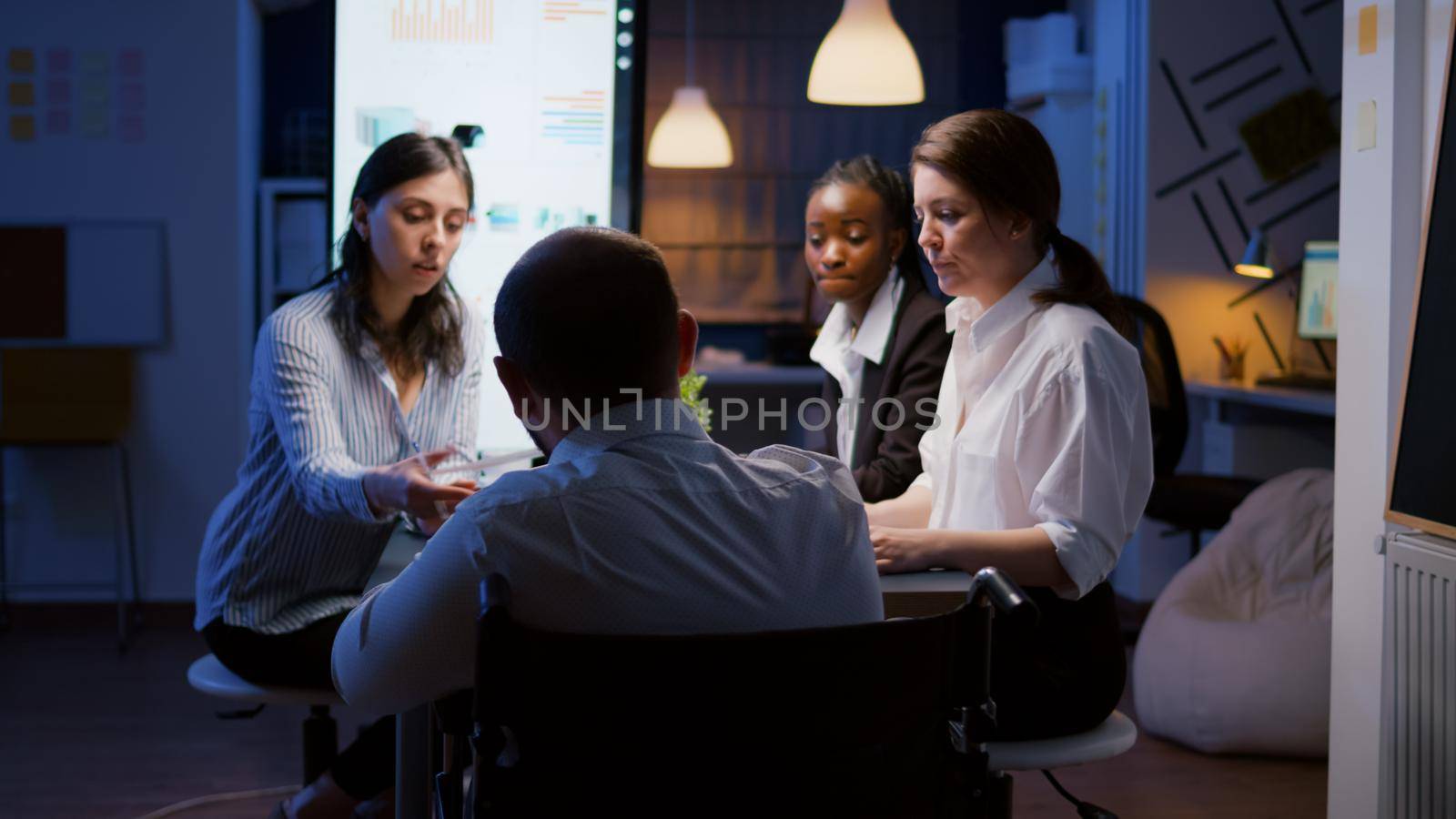 Workaholic disabled businessman in wheelchair sharing statistics paperwork overworking in office meeting room late at night. Diverse of multi-ethnic teamwork analyzing financial strategy