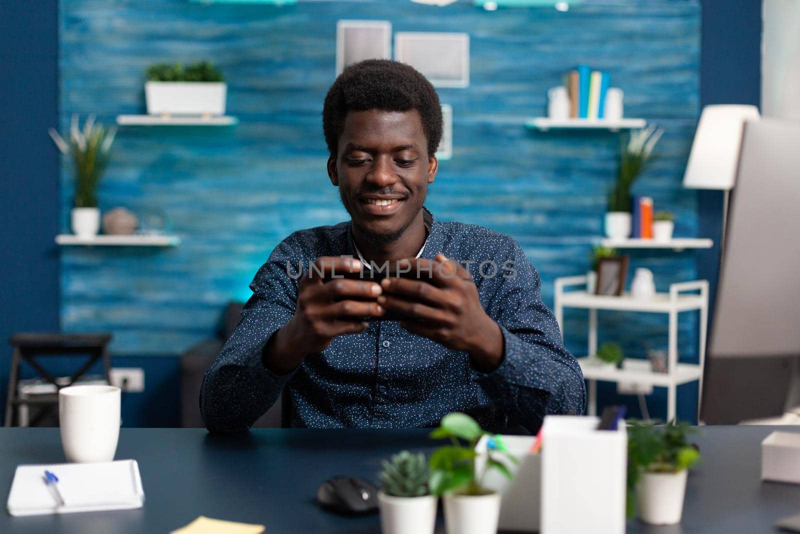 African american man smiling while using a smartphone at desk to check social media. Remote working guy taking a break from assignment, using phone for relaxation and communication