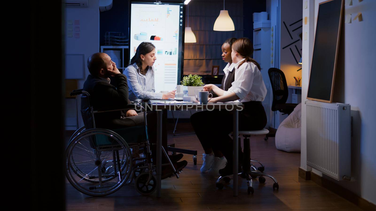 Overworked disabled businessman in wheelchair is ignored while working in company business office meeting room late at night. Workaholics multi-ethnic businesspeople brainstorming strategy ideas