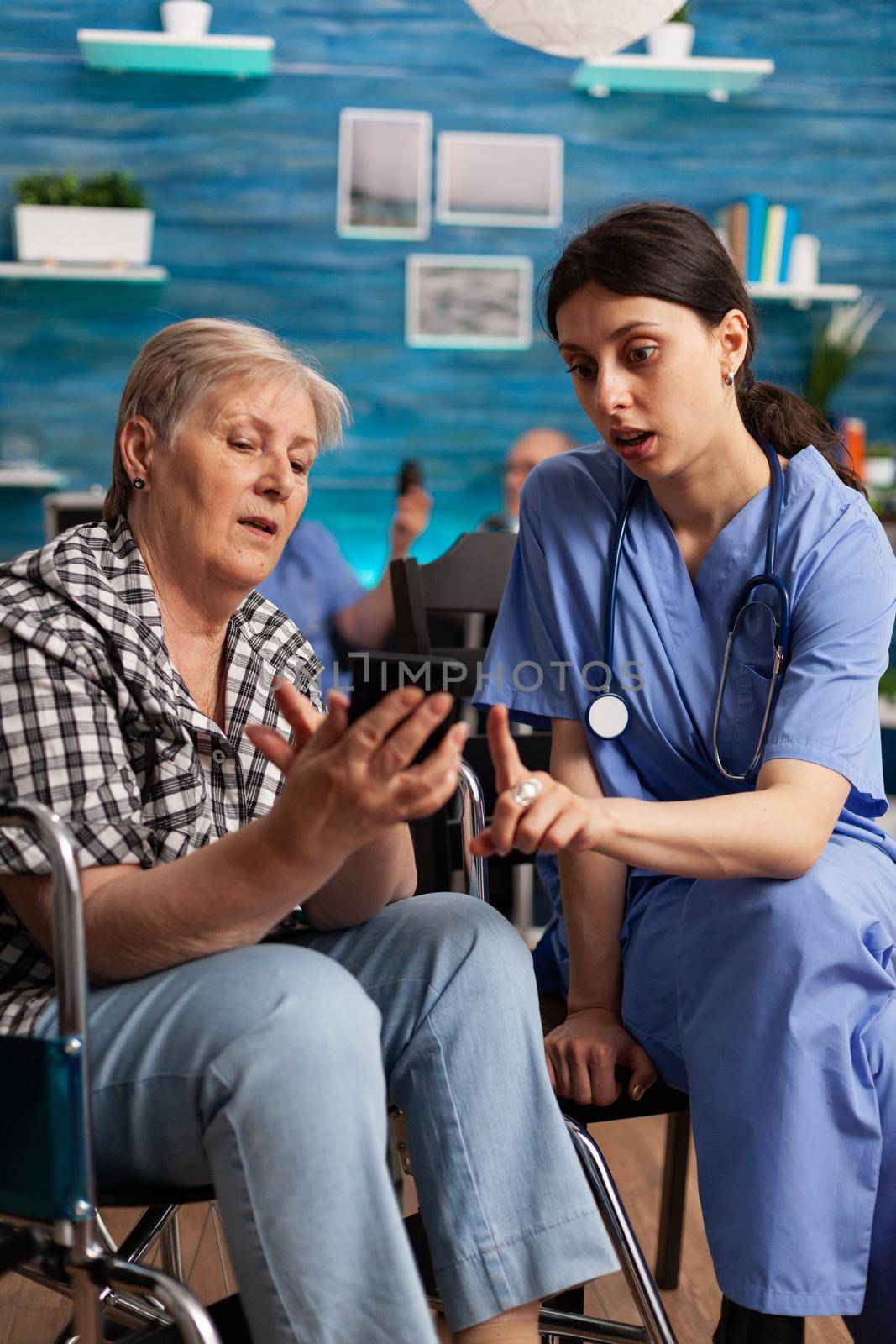 Nurse helping retired senior woman in wheelchair to use smartphone during social service. Elderly person browsing news on digital device in nursing home. Social worker in retirement home