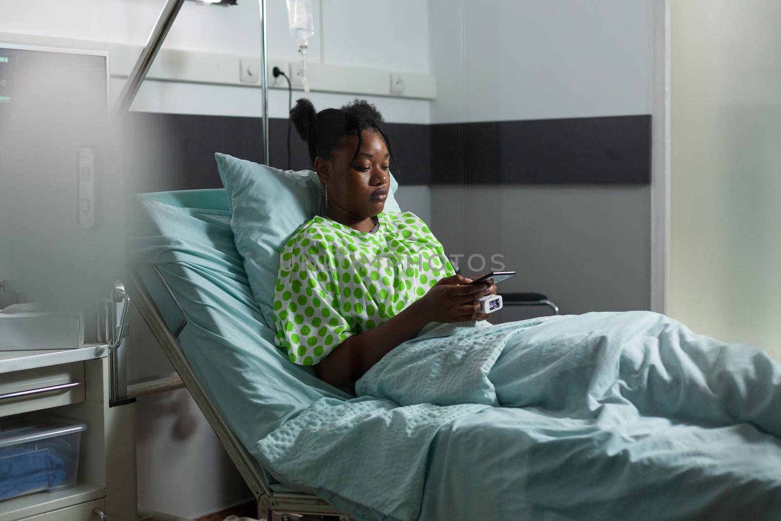 Young african american adult with sickness having smartphone in hand while laying in hospital ward. Sick teenager using internet technology for remote family communication and chatting